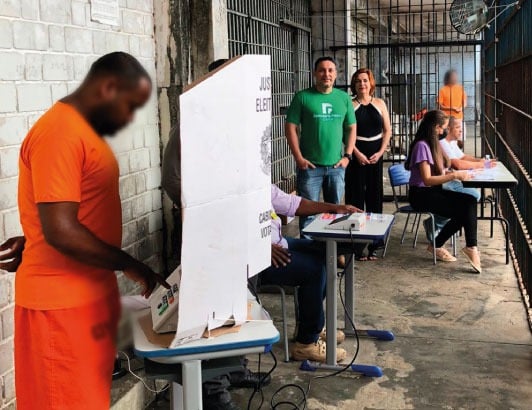 Urna eleitoral brasileira com grade de prisão ao fundo, simbolizando suspensão do voto de presos